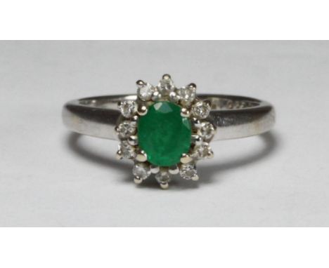 An 18ct white gold ring, centrally claw set with an oval faceted emerald coloured stone, surrounded by twelve small diamonds,