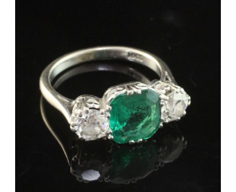 A mid 20th century 18ct white gold, emerald and diamond three stone ring, the central emerald flanked by two round brilliant 