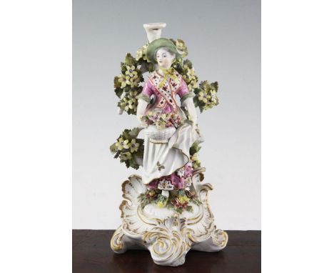A Derby candlestick figure, c.1770, modelled as a lady holding a basket of flowers in her apron standing before bocage, on a 