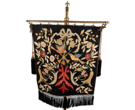 An Antique engraved brass pole stand, on tripod legs and leaf feet, with an embroidered hanging tapestry panel depicting foli