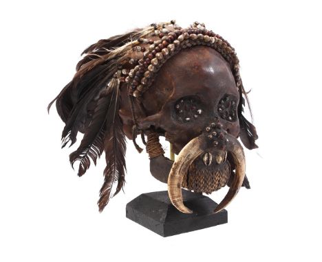 Brown lacquered Asmat skull, decorated with shells, bones and feathers, standing on a blackened wooden base, Papua New Guinea