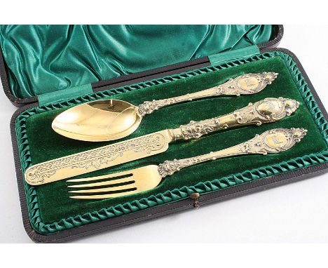 A VICTORIAN ORNATE SILVERGILT CHRISTENING KNIFE, FORK & SPOON crested, in a lined & fitted case, the knife handle loaded, by 