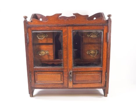 An oak two glazed door smoker's cabinet, opening to reveal two pipe racks and four drawers, with two shaped central reserves,