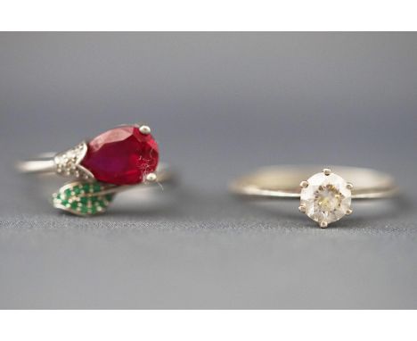 A collection of two white metal rings - One stylised as a crossover flower set with a synthetic ruby and cubic zirconias toge