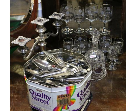 A tin of mixed stainless steel flatwares along with a silver plated candlestick and a collection of glassware.