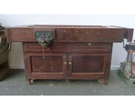 A vintage workbench on casters with two drawers and two cupboard doors beneath, two vices at either end and one to the side t
