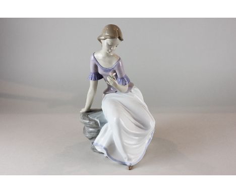 A Nao porcelain figure of a young girl sitting on a rock looking at a flower, 28cm high