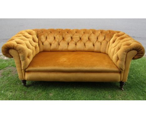 A late Victorian two seat Chesterfield sofa, with rolled arms, buttoned back and mustard coloured upholstery, raised on turne