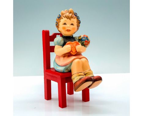 A figure of a young girl in a dress holding a heart shaped container and bouquet of flowers and a red wooden chair she can be