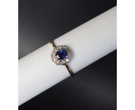 A sapphire and diamond cluster ring set with a central oval faceted sapphire surrounded by round cut diamonds to a white meta