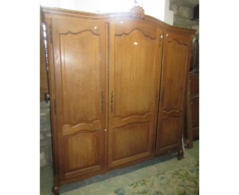 A light oak armoire, with moulded arched pediment over three full length moulded panelled doors, raised on squat scrolled cab