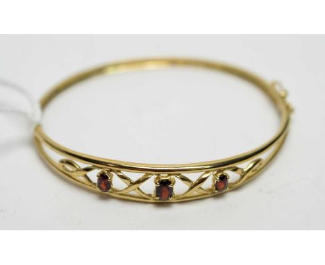 A garnet set yellow metal bangle, the three oval faceted cut graduated garnets in scrolling mount, 6.4cms wide, 8.1g gross.