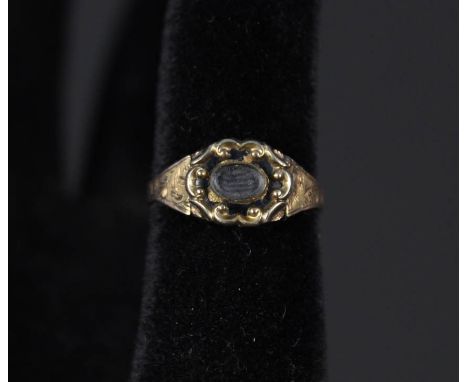 A yellow metal memorial ring with engraved shoulders, black enamel border around the central oval glass panel (some losses to