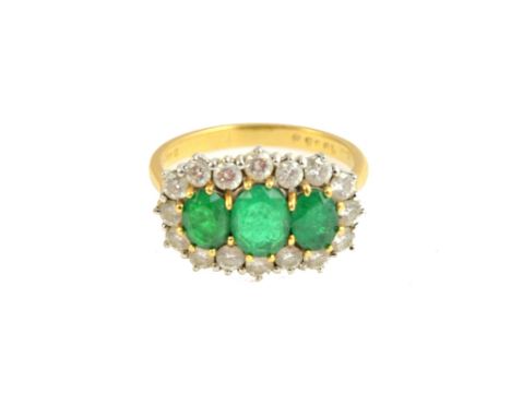 An 18ct gold, emerald and diamond set ring, claw set with a row of three oval cut emeralds at the centre, within a surround o