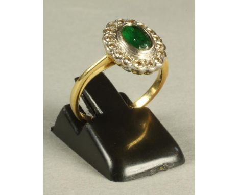 AN EMERALD AND DIAMOND CLUSTER RING, the oval facet cut emerald collet set to a border set with sixteen small diamonds to pla