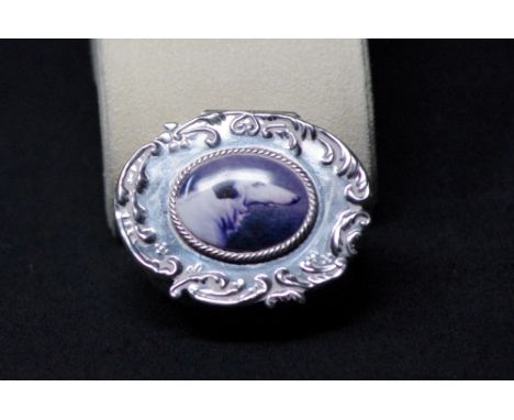 An embossed silver pill box with the lid inlaid with an enamel panel depicting a lurcher dog. Stamped Sterling.