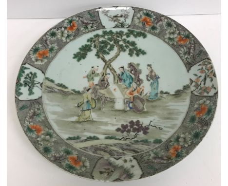 A Kangxi style porcelain charger decorated with figures around a table in a garden setting within a floral and panel decorate