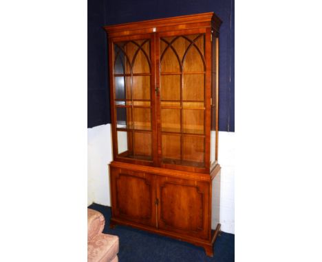 A modern yew wood display cabinet, with two glazed astragal doors enclosing shelved interior, above two panelled doors, endin