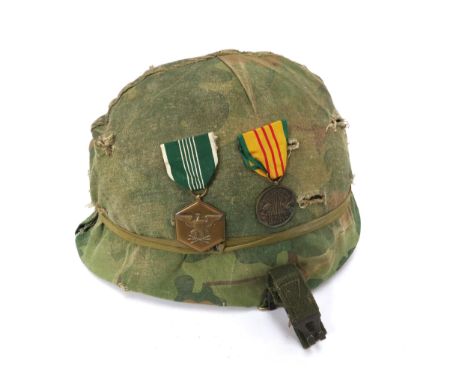 A US Vietnam war era helmet with camouflage cover, the liner dated 1974, with two applied medals