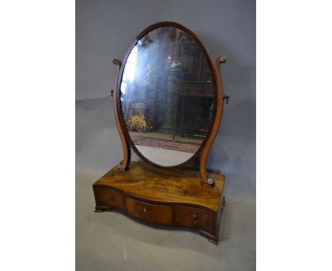 A George III Mahogany and Satinwood Toilet Mirror, the oval mirror with shaped supports above a serpentine fronted box base w