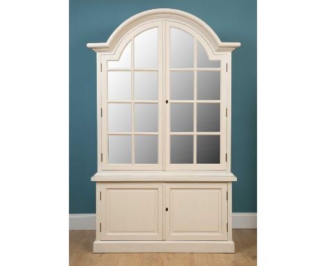 A cream painted OKA display cabinet with an arched top above glass panelled doors, supported by a pair of cupboards raised on