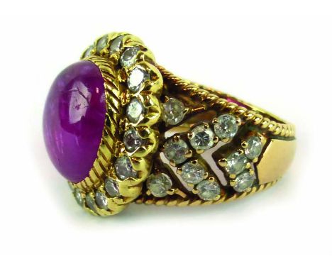 A yellow metal ring set centrally with a cabochon cut oval ruby in a surround of brilliant cut diamonds, the shoulders of che
