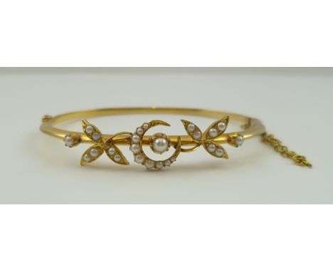 AN 18CT GOLD BANGLE decorated with a seed pearl inset crescent, flanked by seed pearl inset leaves, fitted safety chain, case