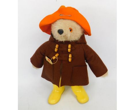 A plush jointed Paddington Bear, with yellow Dunlop type size 4 Wellington boots, brown duffel coat and orange hat, with glas