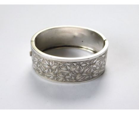 HINGED BANGLE. A broad silver coloured metal hinged bangle, the front engraved with leaves.  Please note that all items in th