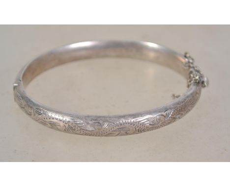 A silver hallmarked bangle bracelet having a half engraved foliate swirl design with safety chain. Weighs 12.6 grams and meas