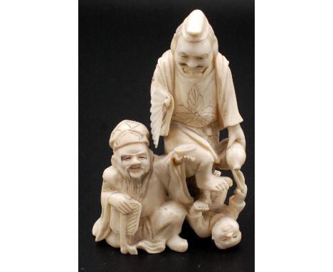 A Japanese ivory okimono, two Sambaso dancers, one standing holding an open fan while playing with an infant lying on it's ba