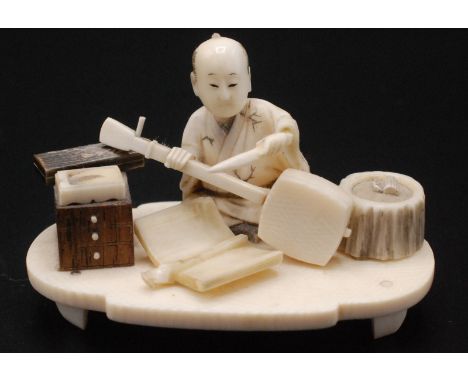 A Japanese ivory okimono, a seated artisan making a samisen, around him are boxes, a three drawer chest, a book, a tool and a