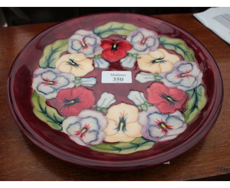 A BURGUNDY GROUND MOORCROFT PLATE decorated with pansies with impressed mark 'Moorcroft' and made in England and diamond stam