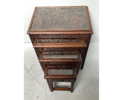 A nest of four Malaysian hardwood tables, each top carved with scenes of figures, buildings and trees, largest measures 51cm 