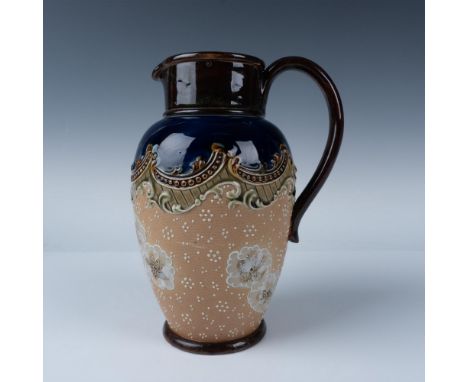 A round pitcher with a light brown body and a blue ring top, decorated with white low relief patterns of dots and flowers. Ro