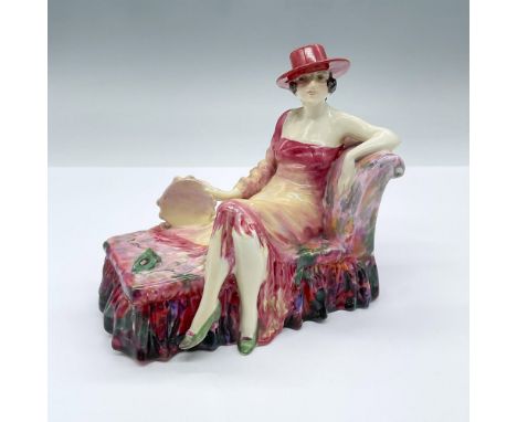 Features a woman seated on a colorful sofa, fashioned in pink, holding a tambourine. Her green shoes match the masquerade mas