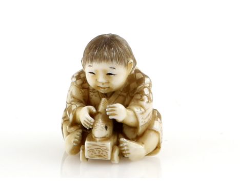 A stained ivory netsuke of a seated boy, playing with a fish toy on wheels, signed KogyokuProvenance: The Property of a Lady.