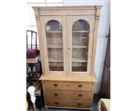 A 19TH CENTURY STRIPPED PINE BOOKCASE or cabinet, with arched glazed doors above three long drawers 197cm high, 105cm wide