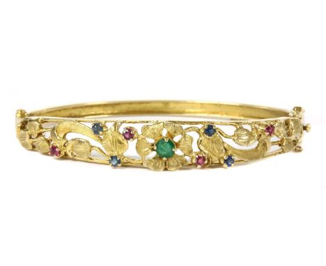 A 9ct gold emerald, ruby and sapphire set hinged bangle, c.1970, with a pierced foliate top half. A circular mixed cut emeral