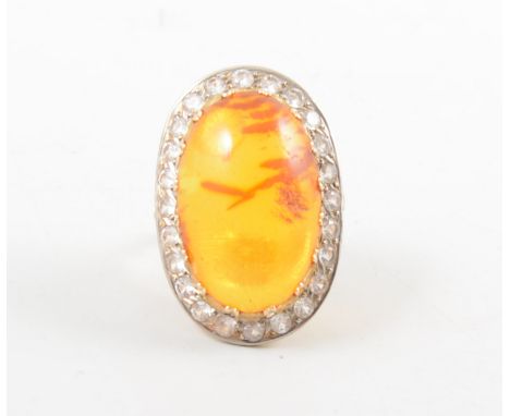 An amber dress ring, the oval cabochon cut amber stone, 25mm x 18mm, claw set and surrounded by twenty-four synthetic white s