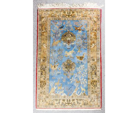 A Qum rug woven in fawn, ivory and pale blue with various animals, birds and flowering trees, on a pale blue ground, within f