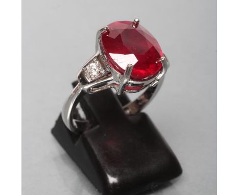 A RUBY RING, the oval facet cut glass filled stone of approximately 7.7cts, claw set to shaped shoulders each set with a bril