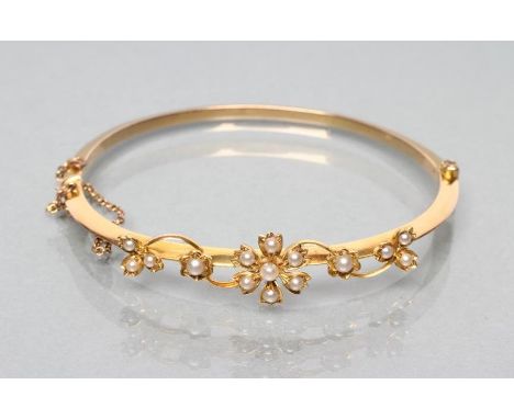 AN EDWARDIAN STIFF HINGED BANGLE, the upper section with open scrolls and seed pearl set flowerhead and leaves, stamped 15ct 