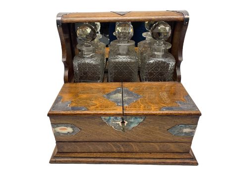 Edwardian oak three-bottle tantalus with silver-plated strapwork mounts, shield shaped plaque and swing handles, containing t