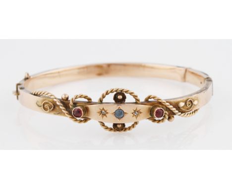 A Victorian 9ct yellow gold hinged bangle, set with central round cut sapphire, flanked with round cut diamonds and red paste