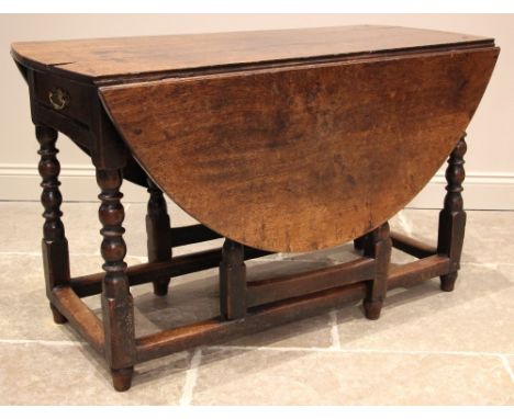 An 18th century oak drop leaf table, the oval plank top over a single frieze drawer, upon bobbin and block supports united by