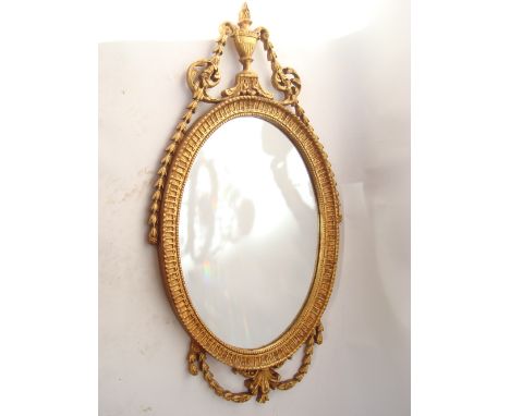 A 20th Century Neoclassical Adam style giltwood and gesso oval wall mirror surmounted by an urn and trailing bell husk traili