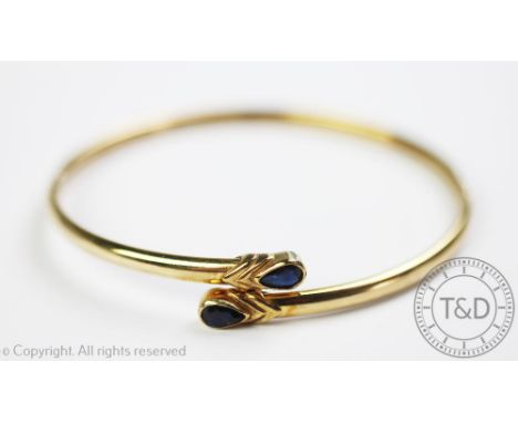 A yellow gold and sapphire set bangle, in cross-over design and each set with a pear shaped sapphire within rub-over collet s