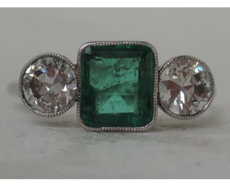 A ladies three stone  dress ring having a central trap cut emerald flanked by two diamonds each approx .5ct in millgrain moun