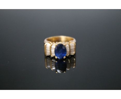 AN 18K SAPPHIRE AND DIAMOND RING, the central oval sapphire measuring approx 9mm by 6mm has baguette cut diamonds set either 